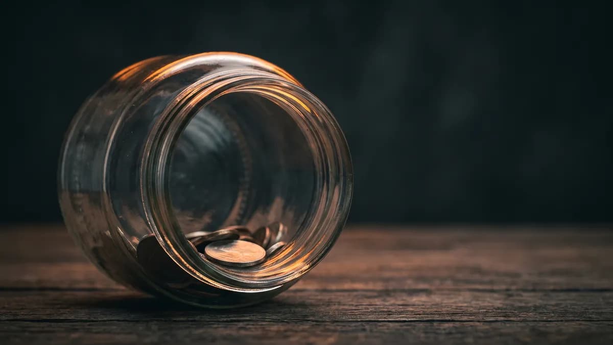 A single glass savings jar holding only a few coins on a bare wooden table, warm amber rim light against a dark background, cinematic photorealism, shallow depth of field